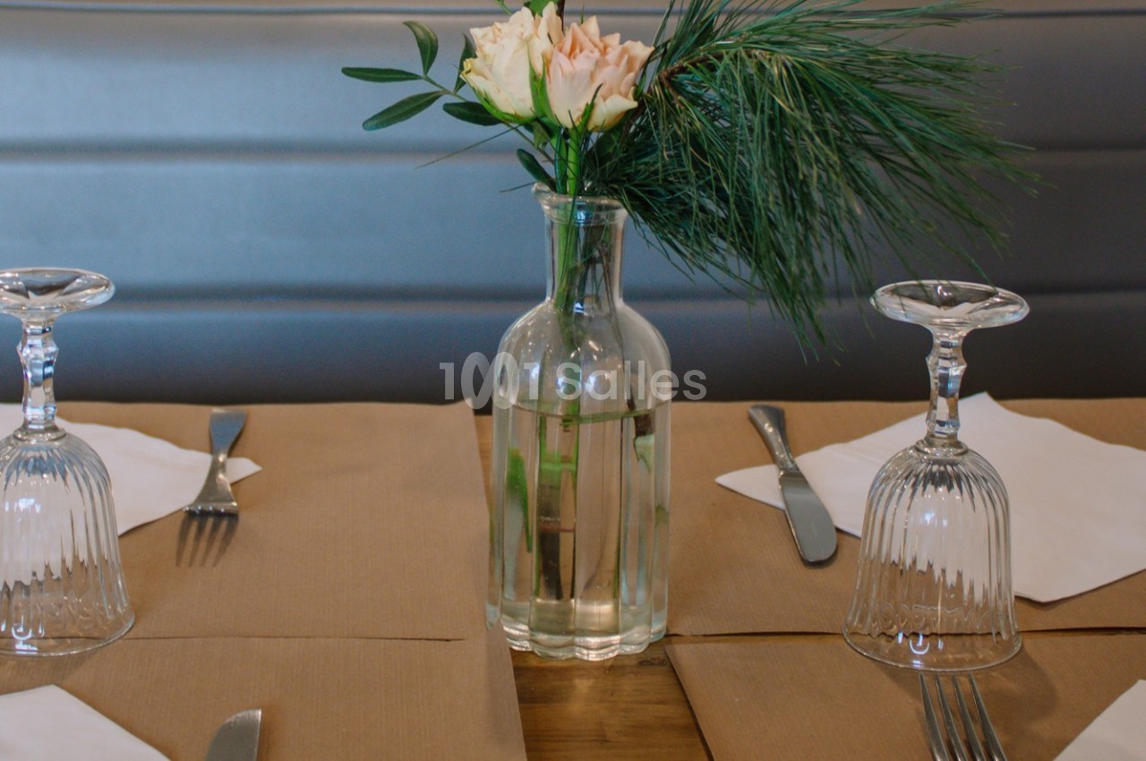 Vase en verre avec des fleurs et des branches vertes, posé sur une table dressée avec des verres et des couverts.