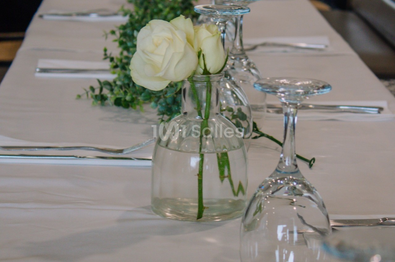 Vase en verre contenant deux roses blanches, posé sur une table dressée avec des verres et des couverts.