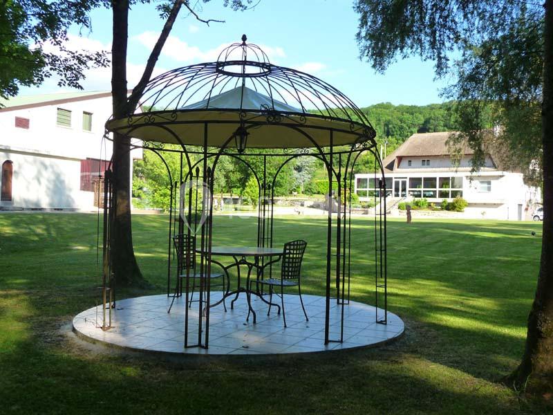 Tonnelle en fer forgé avec table et chaises sur une pelouse, entourée d'arbres et de bâtiments en arrière-plan.