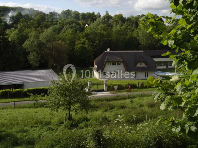 Maison au toit de chaume entourée de verdure, avec un chemin et des arbres en arrière-plan.