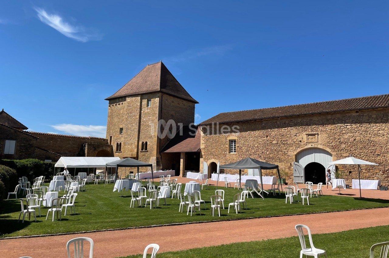 Chaises blanches et tables disposées sur une pelouse devant un bâtiment en pierre sous un ciel bleu.