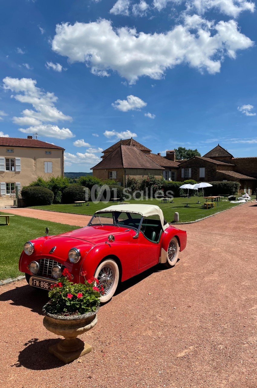 Voiture ancienne rouge décapotable stationnée devant un bâtiment en pierre avec pelouse et ciel dégagé.