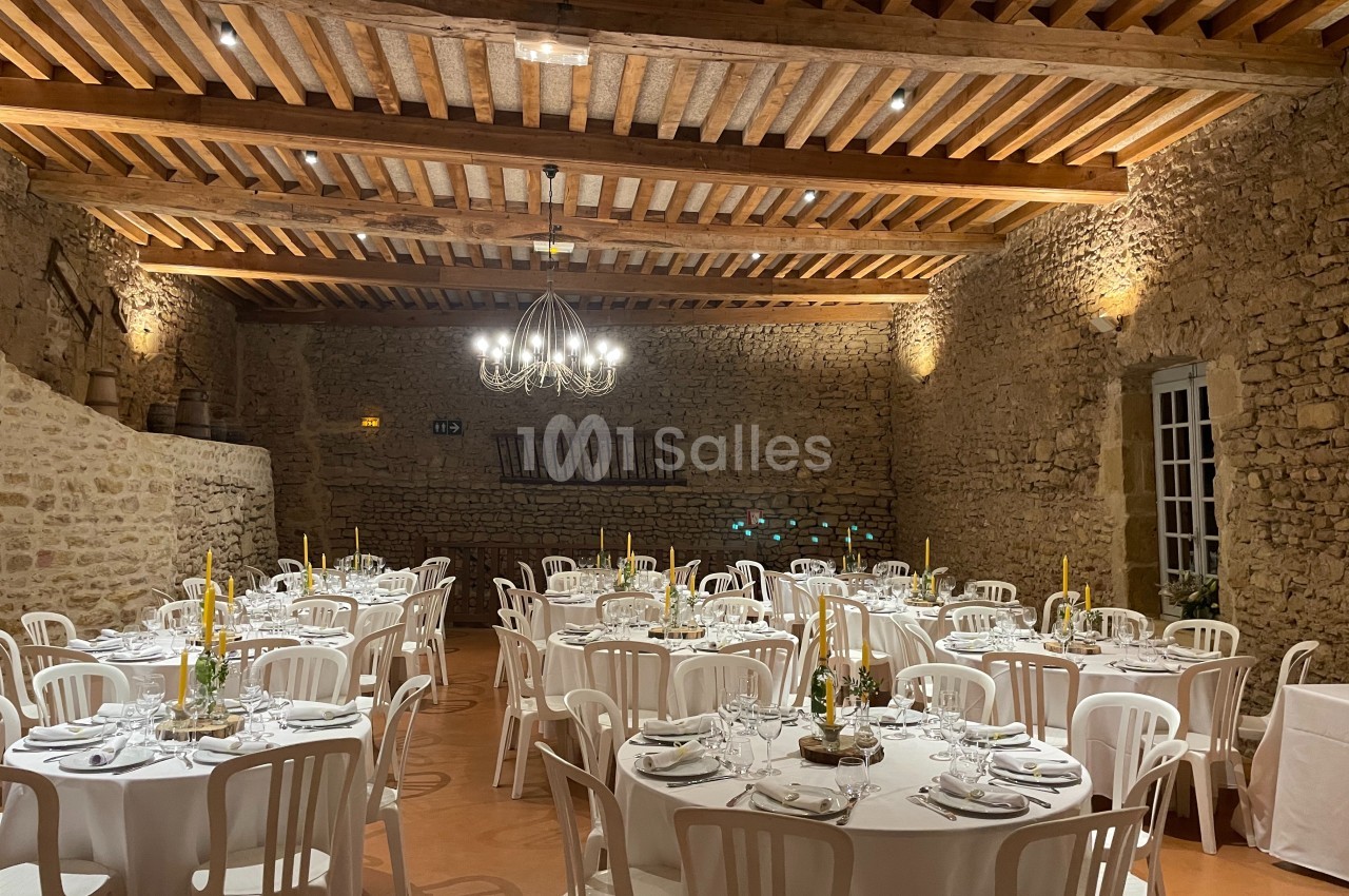 Salle en pierre avec plafond en poutres apparentes, tables rondes dressées avec nappes blanches et chandeliers jaunes.