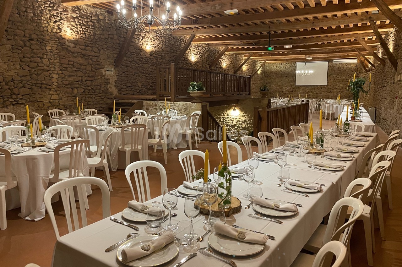 Salle de réception avec murs en pierre, tables dressées avec nappes blanches, chaises en plastique et décoration sobre.