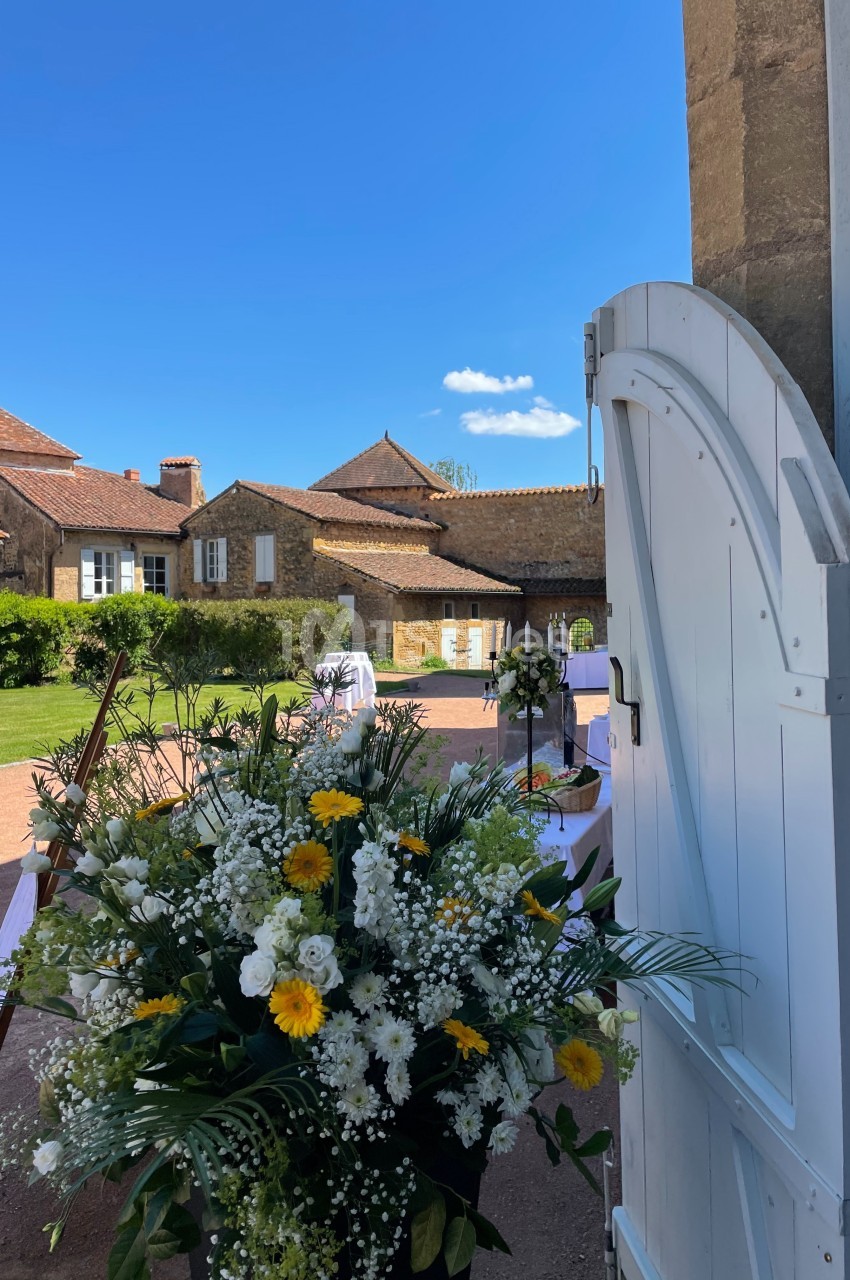 Vue d'une cour ensoleillée avec des bâtiments en pierre, des tables dressées et un bouquet de fleurs blanches et jaunes au…