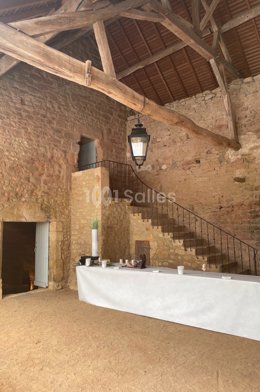 Intérieur d'une grange en pierre avec une charpente en bois, un escalier en fer forgé et une table recouverte d'une nappe…