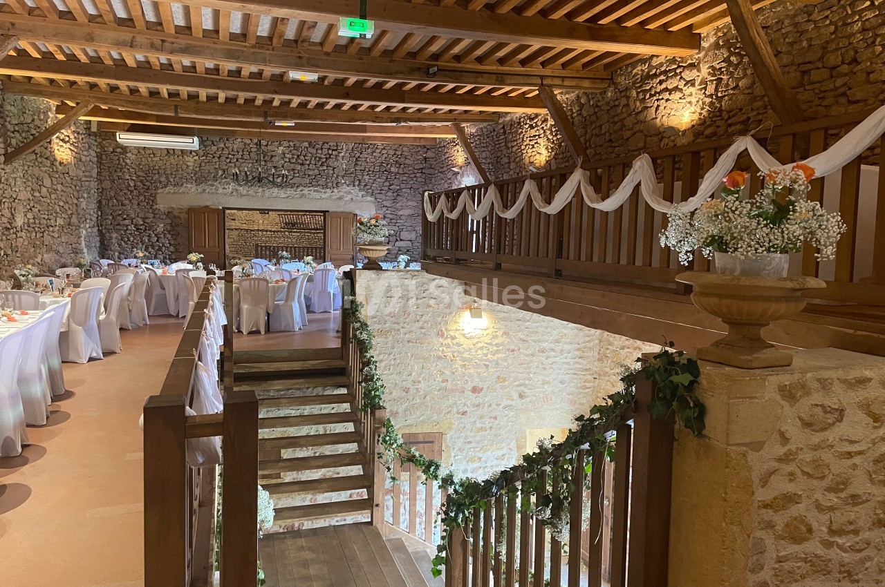 Salle de réception en pierre avec tables dressées, décorations florales et mezzanine en bois ornée de guirlandes.
