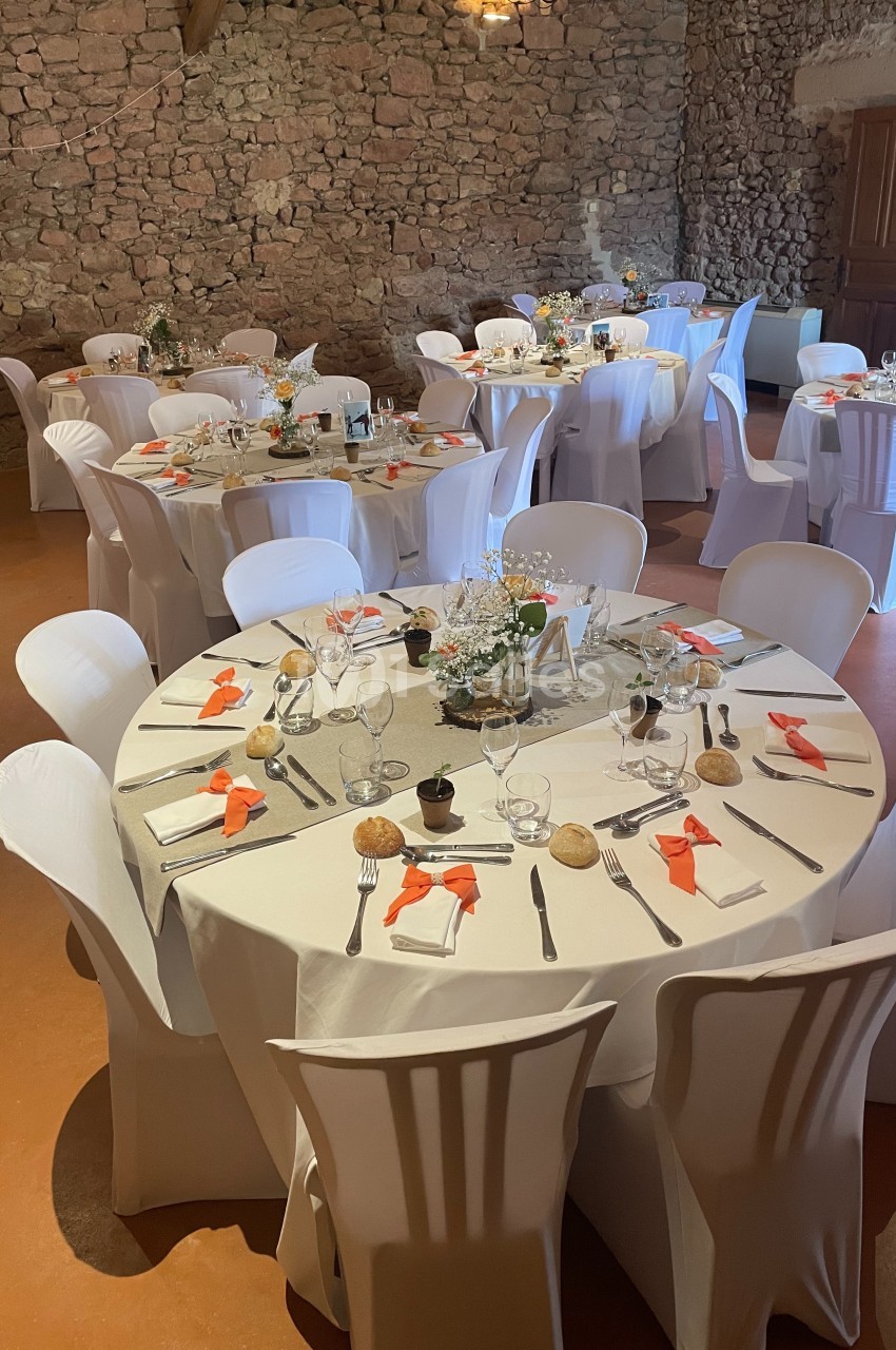 Salle décorée pour un repas, avec des tables rondes dressées, nappes blanches et touches de décoration orange.