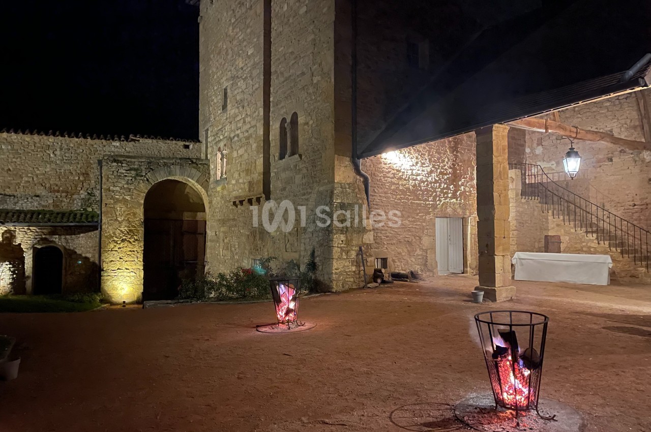 Cour d'un bâtiment en pierre éclairée la nuit, avec deux braseros allumés et un escalier en arrière-plan.