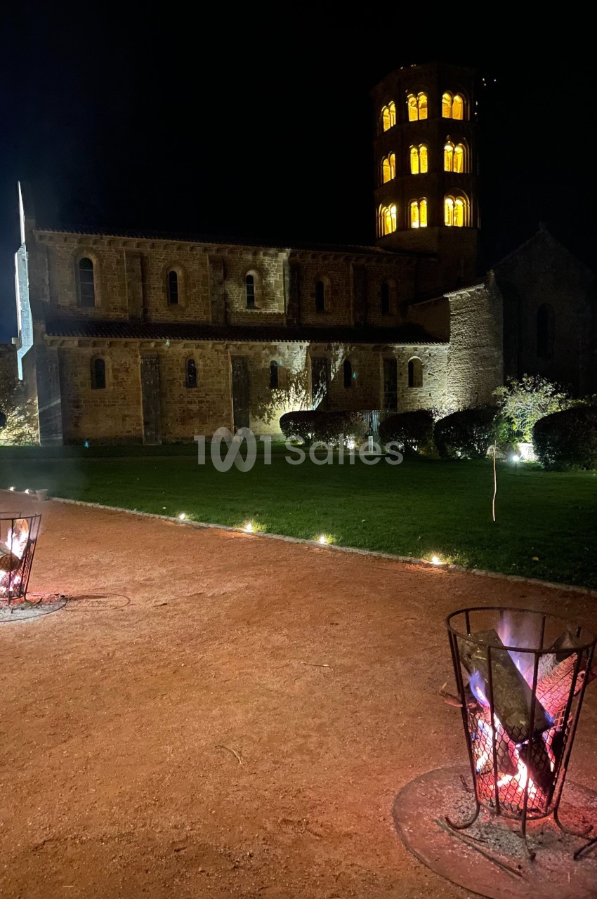 Église illuminée de nuit avec un clocher éclairé, vue depuis une allée avec des braseros allumés au premier plan.