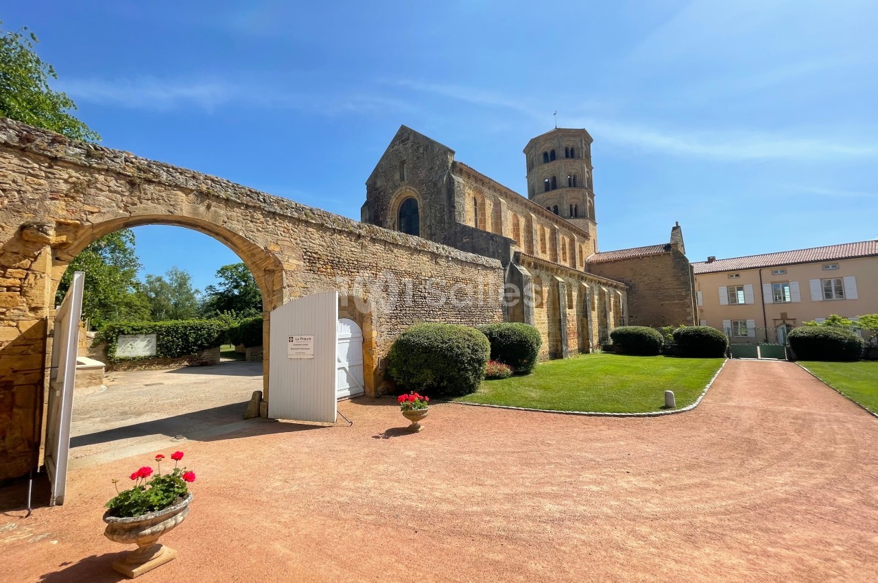 Vue d'une église romane en pierre avec un clocher, entourée d'un jardin et d'une allée en gravier sous un ciel bleu.