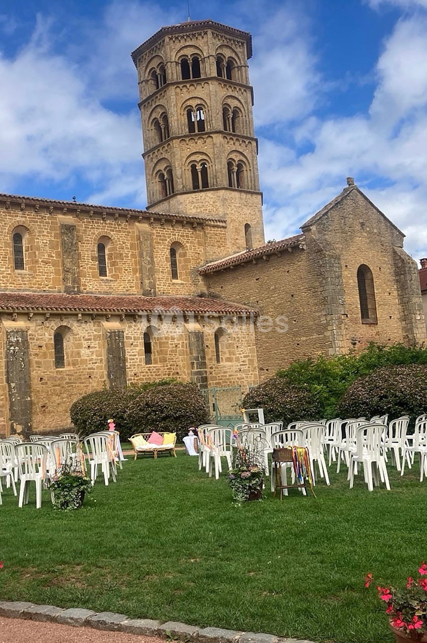 Église en pierre avec clocher roman, entourée de chaises blanches disposées sur une pelouse sous un ciel partiellement…