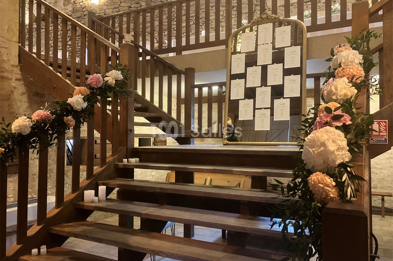 Escalier en bois décoré de fleurs et bougies, avec un tableau de plan de table placé au centre dans une salle en pierre.