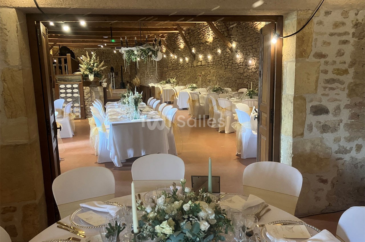 Salle de réception en pierre avec tables décorées de nappes blanches, chaises ornées de rubans et centre de table floral.