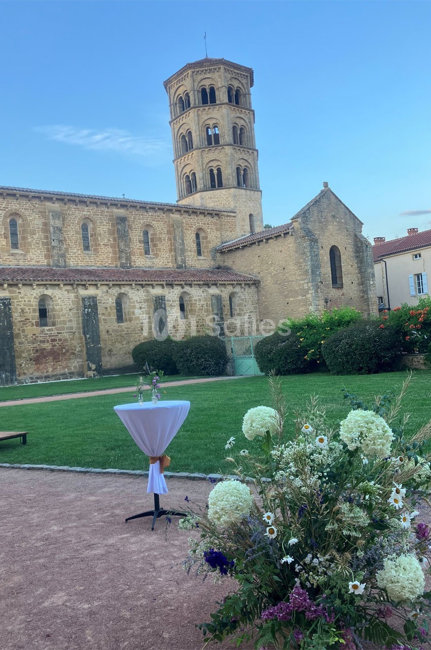Vue d'une église romane avec un clocher, un jardin fleuri et une table haute décorée au premier plan.