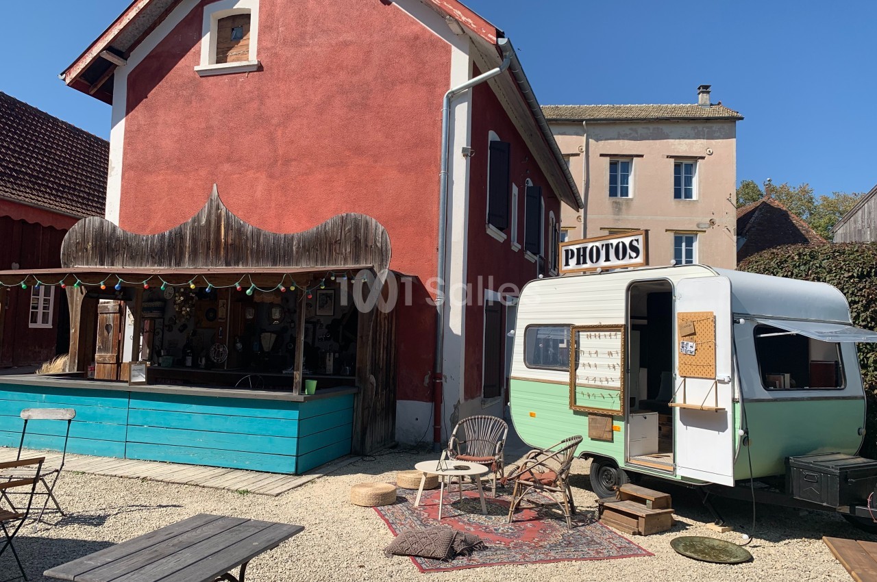 Façade d'une maison rouge avec un bar en bois et une caravane aménagée en stand photo dans une cour ensoleillée.