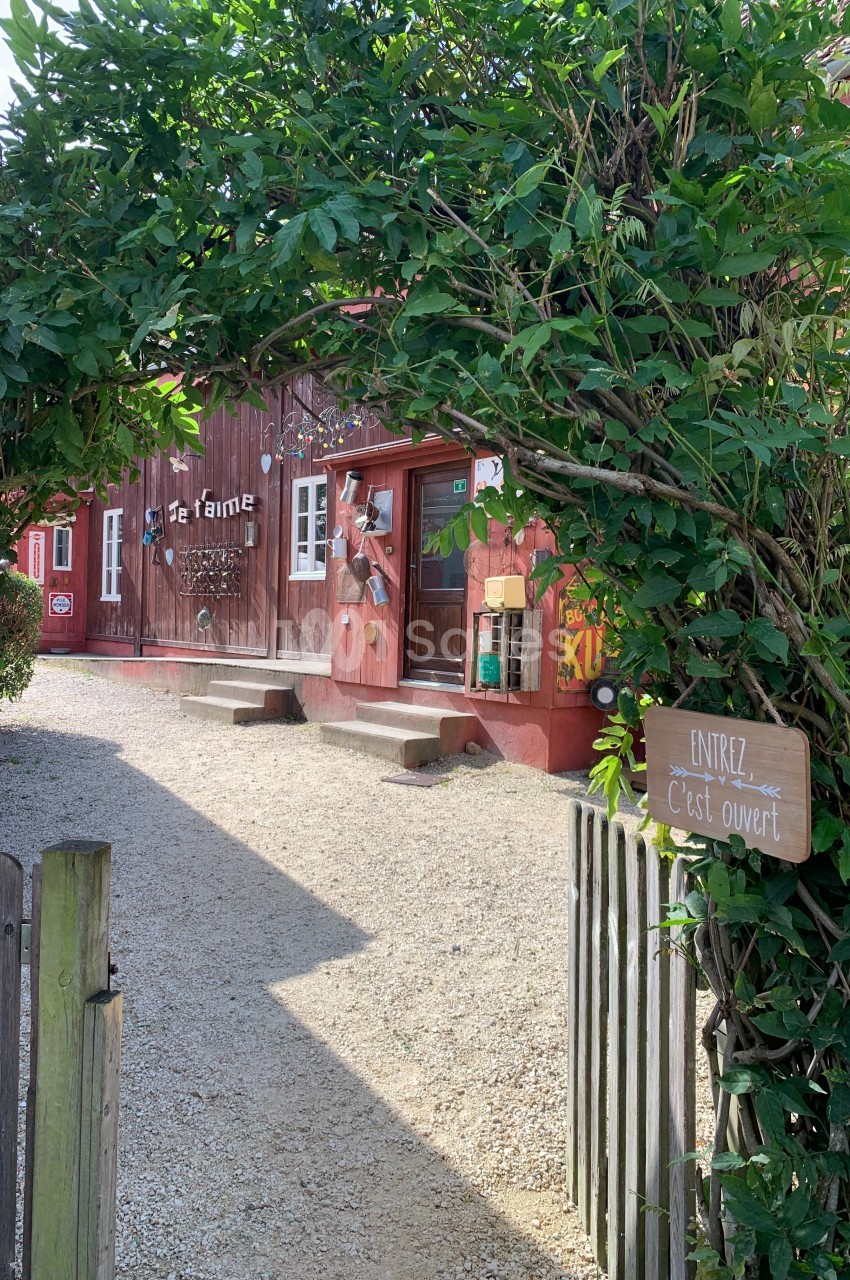 Entrée d'une ferme avec un portail entouré de verdure, un panneau ’Entrez, c'est ouvert’ et un bâtiment en bois rouge.