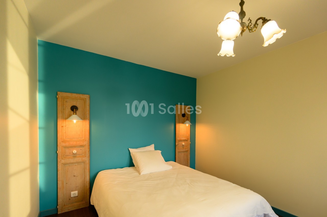 Chambre avec un lit simple, mur bleu, deux appliques murales et un plafonnier à trois abat-jours blancs.
