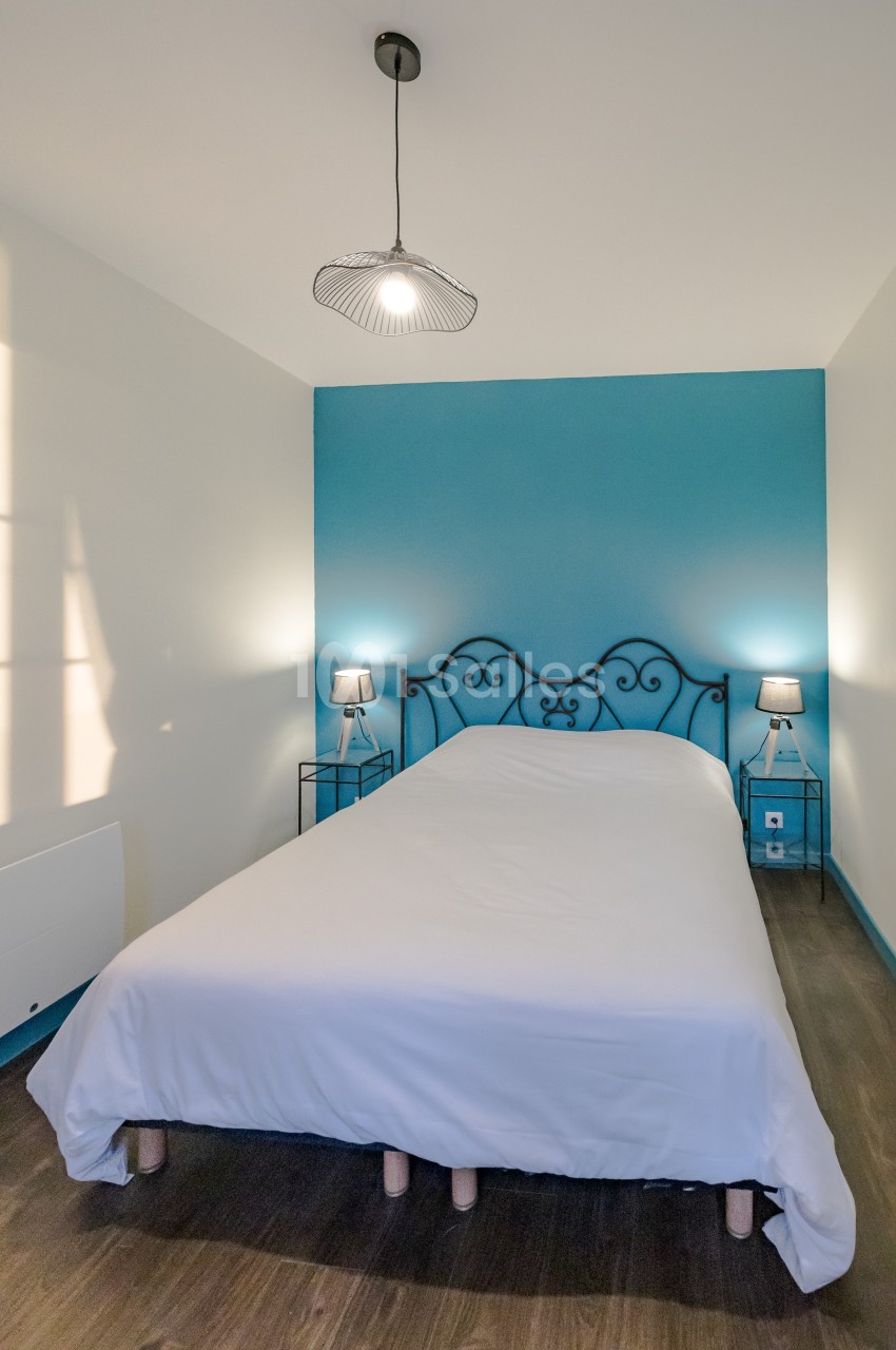 Lit double avec drap blanc, encadré par deux tables de chevet et lampes, dans une chambre au mur bleu et sol en bois.