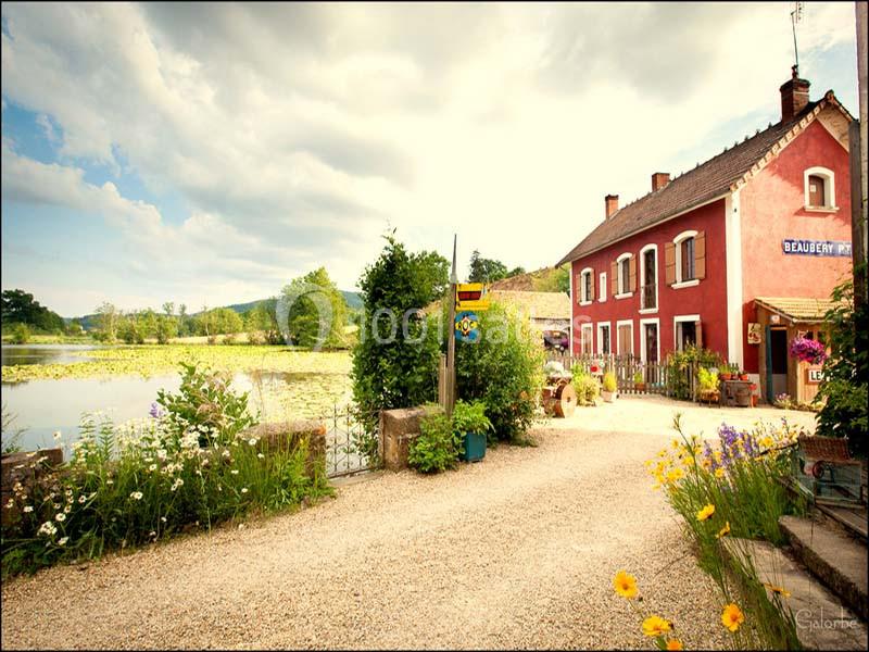 Chemin bordé de fleurs menant à une maison rouge près d'un étang sous un ciel partiellement nuageux.
