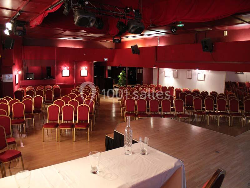 Salle de conférence avec des rangées de chaises rouges, une scène en bois et une table couverte d'une nappe blanche.