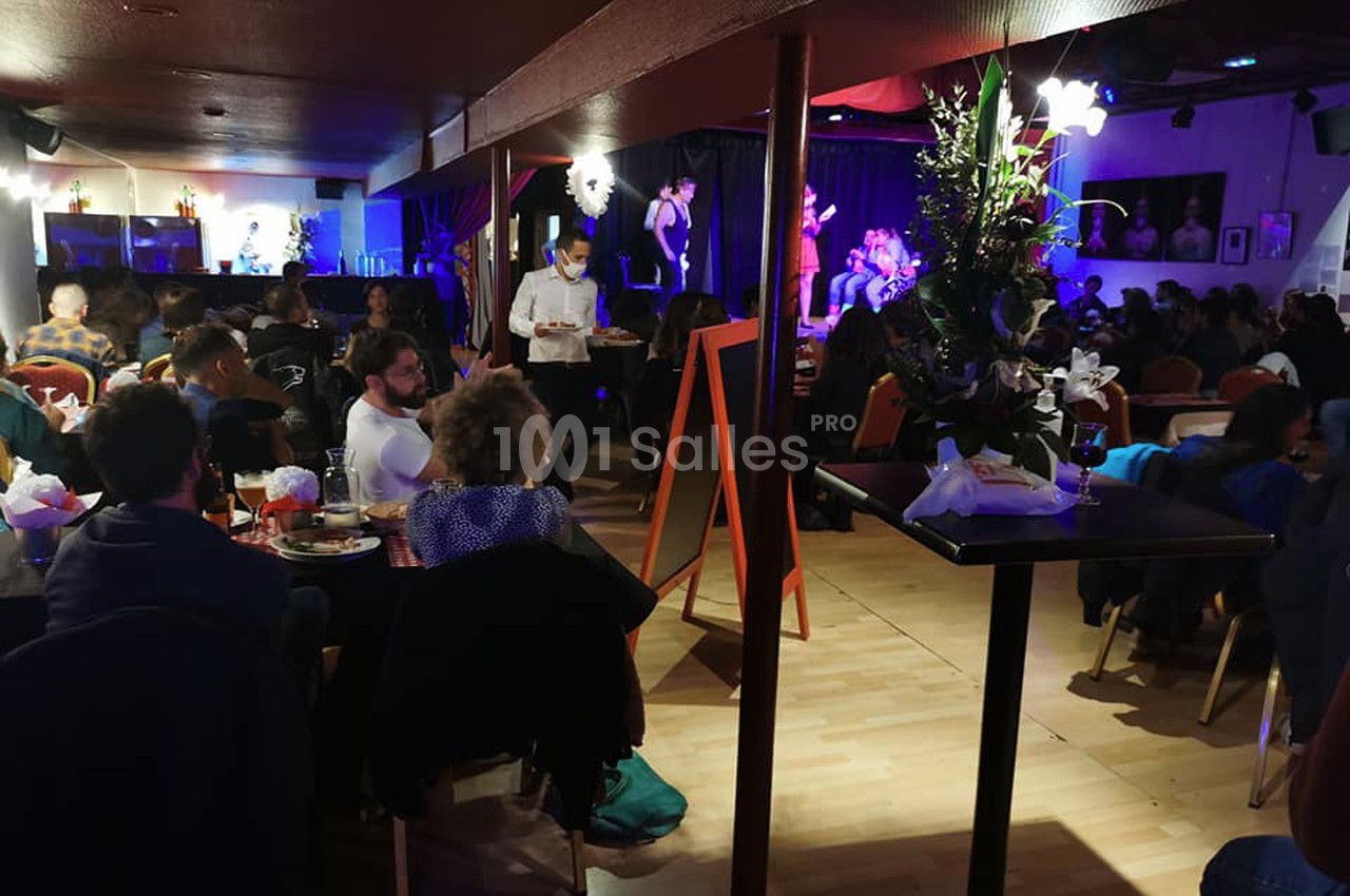 Salle animée avec des tables occupées, un serveur en mouvement et une scène où des musiciens jouent sous des lumières…