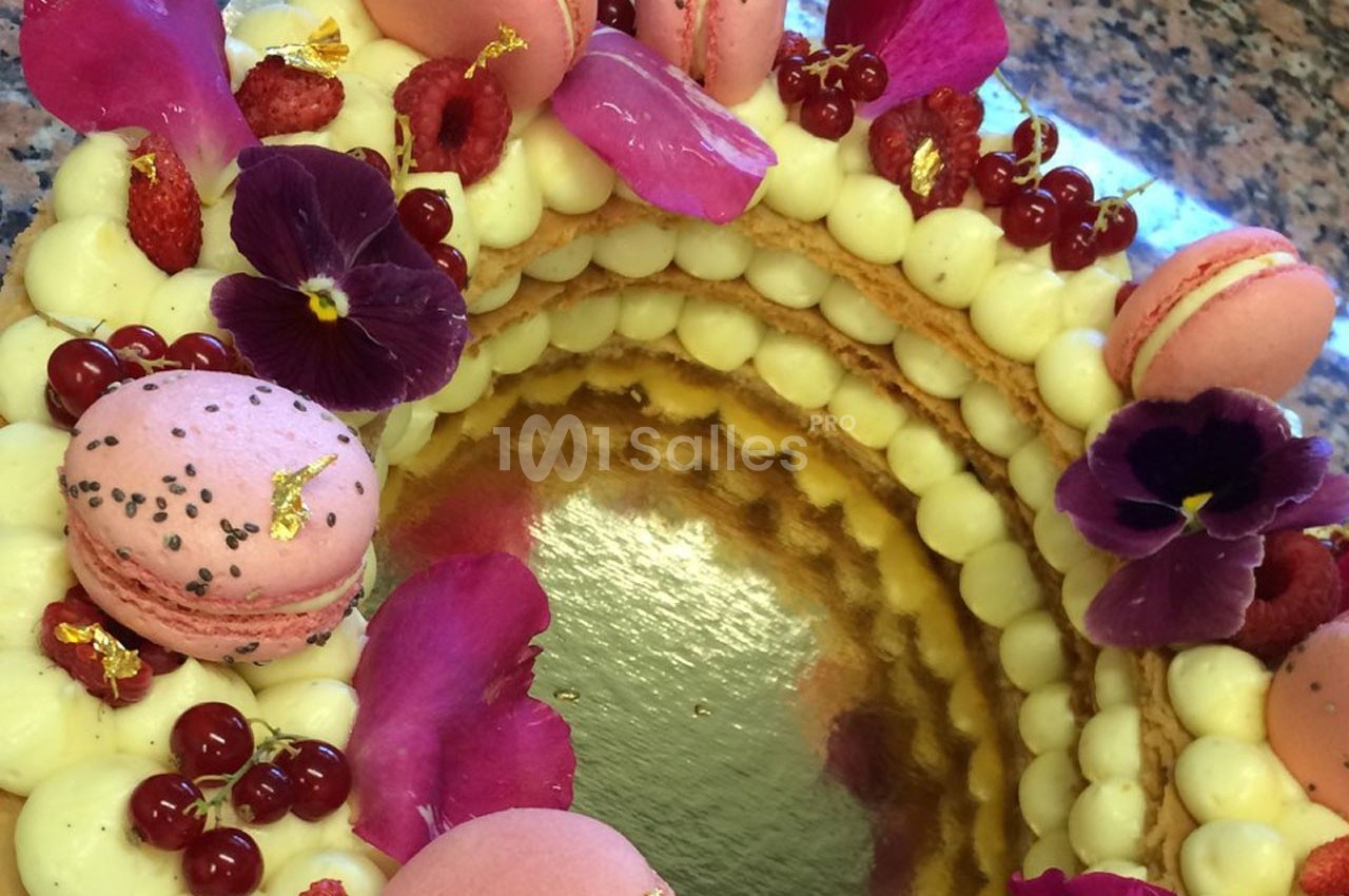 Gâteau décoré de macarons roses, fleurs comestibles, framboises et groseilles rouges sur une base de crème.