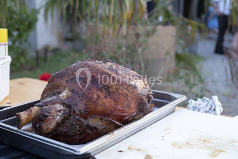 Gigot d'agneau rôti posé dans un plat en métal sur une table en extérieur, avec un fond flou de jardin.
