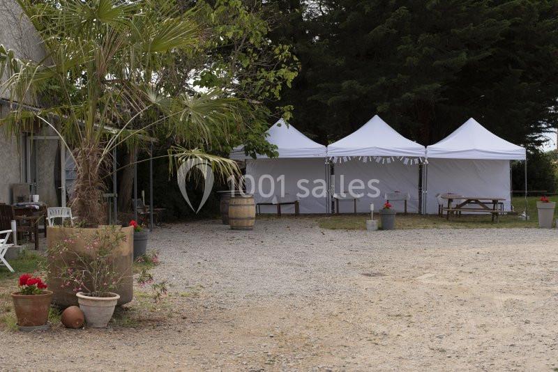 Trois tentes blanches installées sur un sol gravillonné près d'arbres, avec des tables et des plantes en pot.