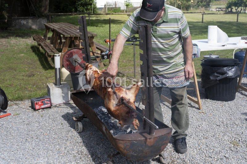Homme faisant rôtir un cochon entier sur une broche au-dessus de braises dans un espace extérieur.