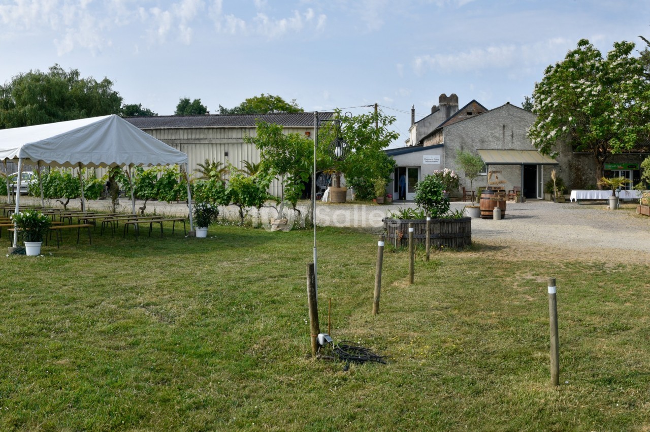 Vue d'un espace extérieur avec une tente blanche, des tables, des chaises et des bâtiments entourés de verdure.