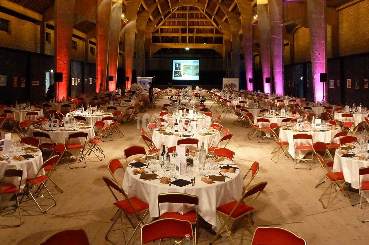 Salle de réception avec de grandes tables rondes dressées, chaises rouges et éclairage d'ambiance coloré.