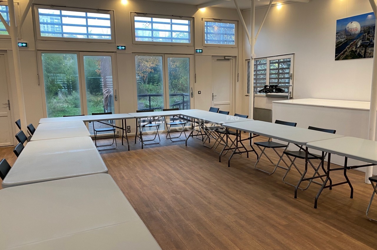 Salle lumineuse avec tables en U, chaises pliantes, parquet au sol et grandes baies vitrées donnant sur un espace vert.