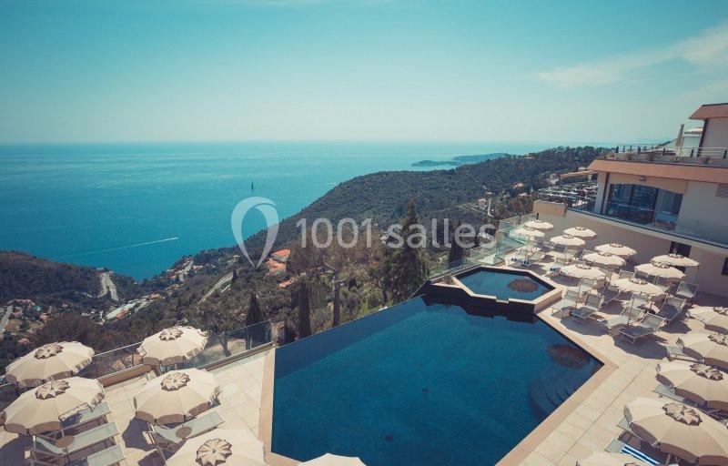 Vue d'une piscine extérieure surplombant une colline verdoyante et la mer, entourée de parasols et de chaises longues.