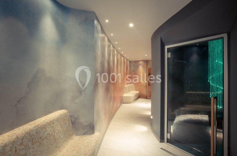 Couloir d'un spa avec éclairage tamisé, murs texturés et bancs en mosaïque, menant à une porte en verre.