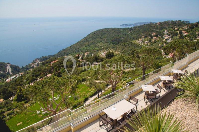 Vue sur une terrasse avec tables et chaises, surplombant des collines verdoyantes et la mer Méditerranée au loin.