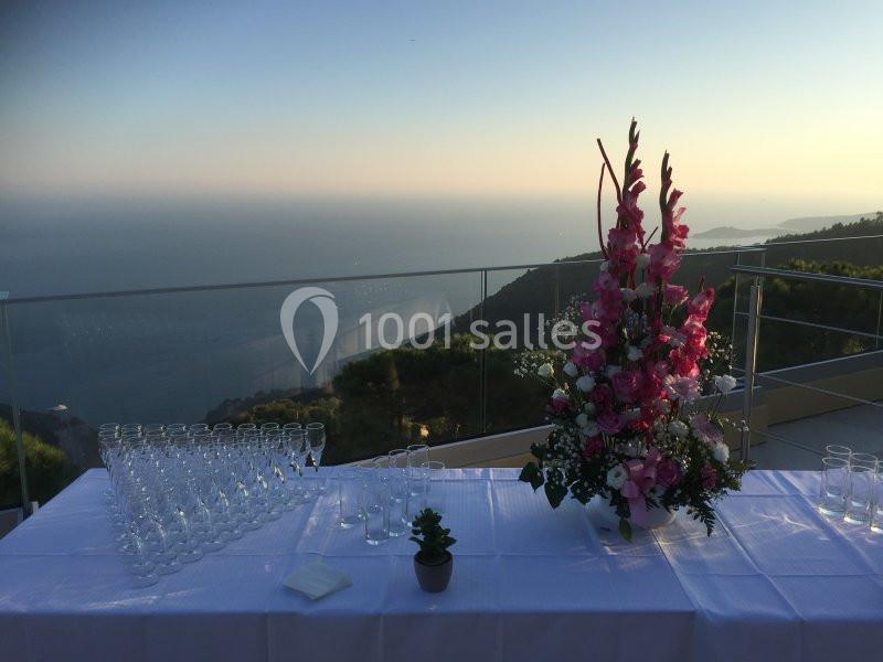 Table dressée avec des flûtes à champagne et un grand bouquet de fleurs roses, en terrasse avec vue sur la mer au coucher du…