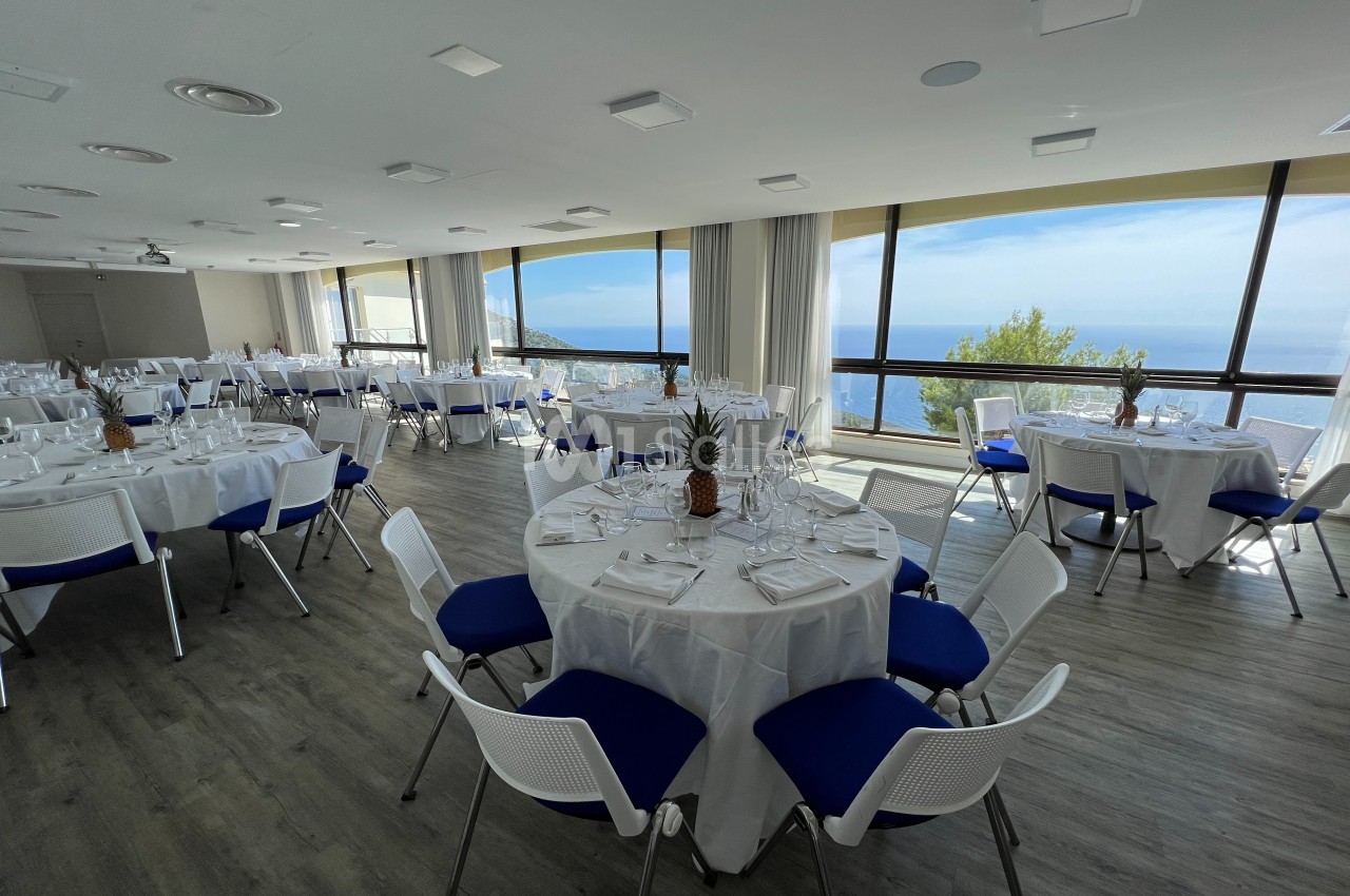 Salle lumineuse avec tables rondes dressées, nappes blanches, chaises bleues et vue panoramique sur la mer.