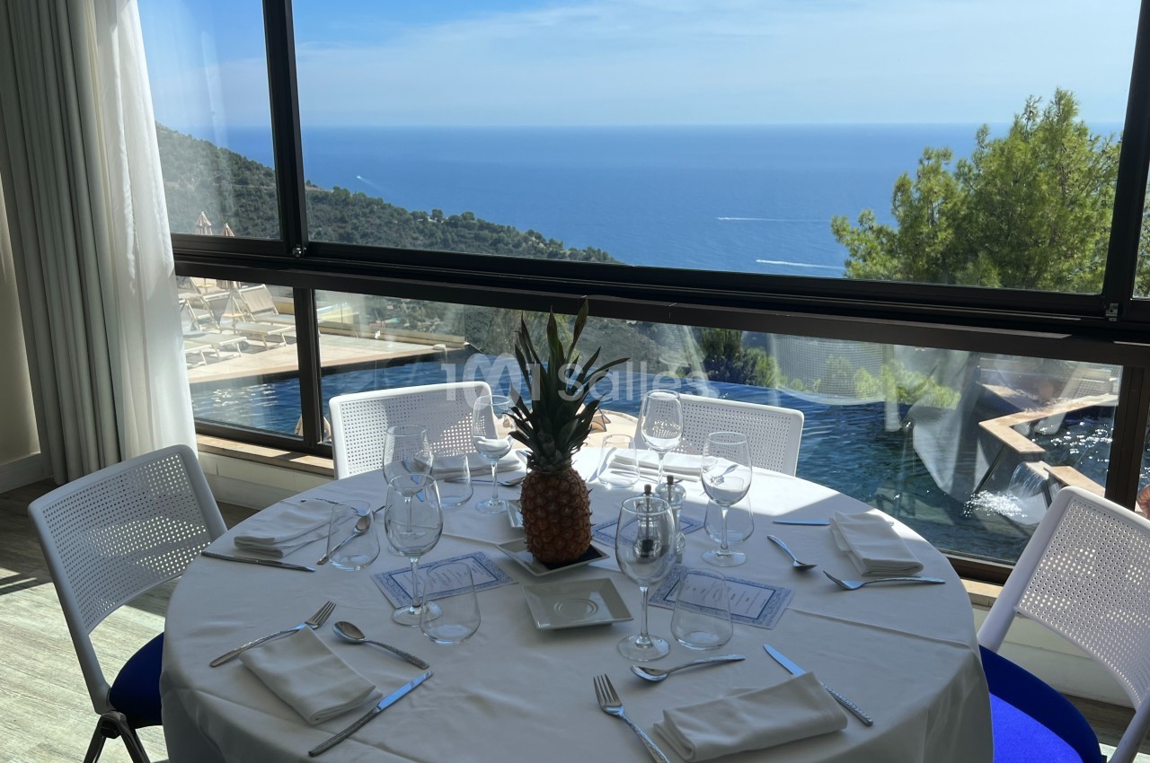 Table dressée avec vue panoramique sur la mer, entourée de baies vitrées et d'une piscine extérieure.