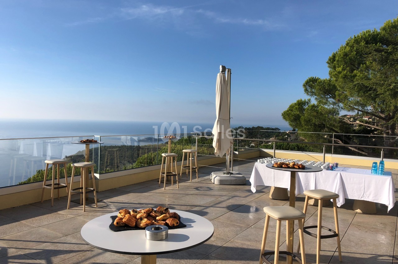 Terrasse ensoleillée avec vue sur la mer, tables hautes, viennoiseries et boissons disposées pour un buffet.