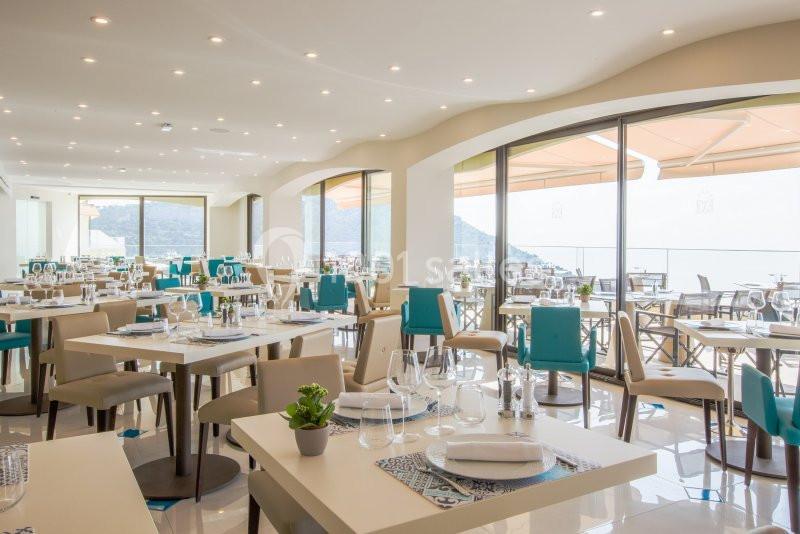 Salle lumineuse d'un restaurant avec tables dressées, grandes baies vitrées et vue sur un paysage côtier.