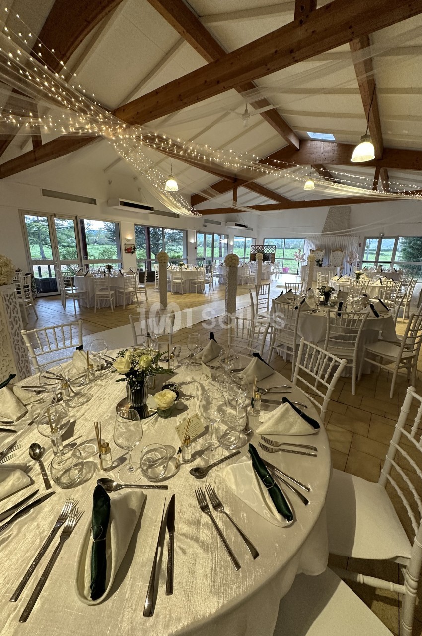 Salle de réception lumineuse avec tables élégamment dressées, chaises blanches et décorations suspendues.