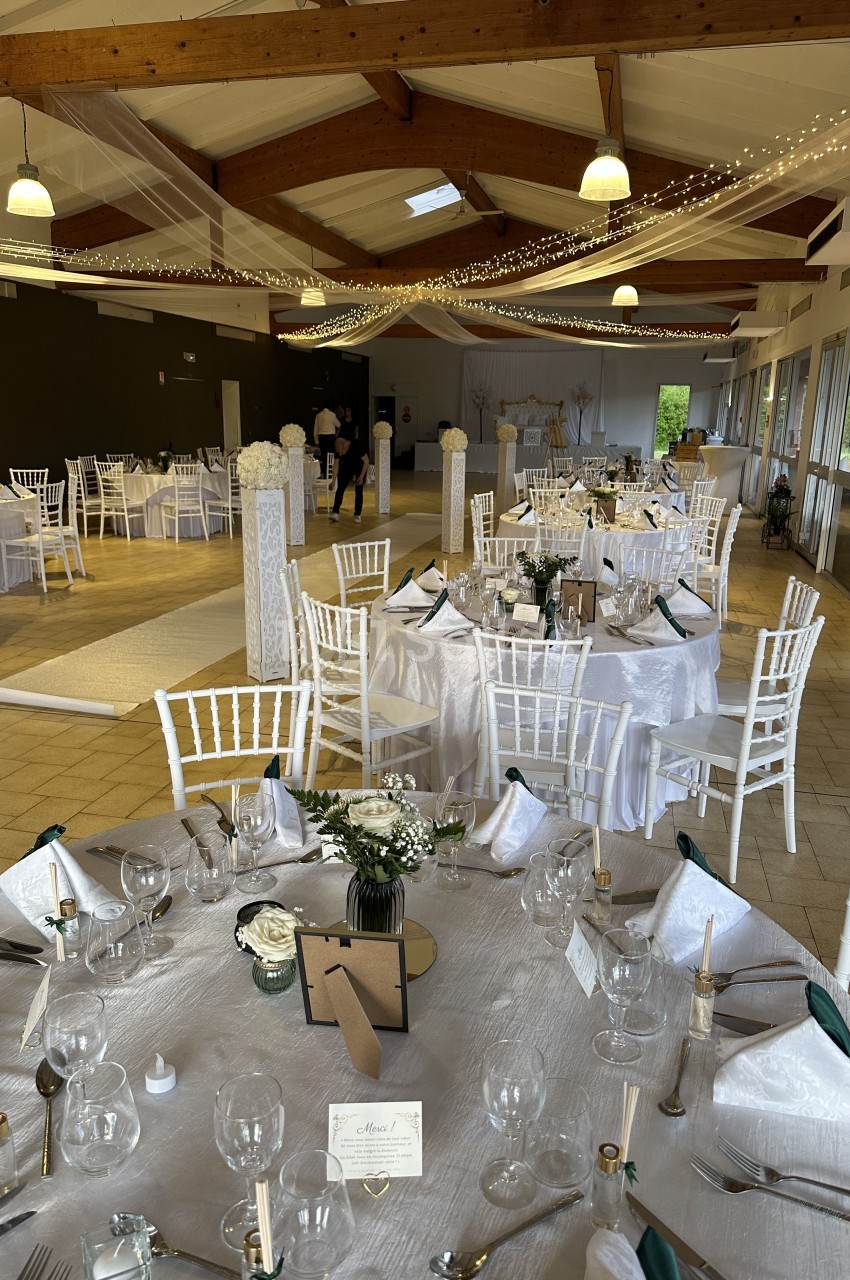 Salle de réception décorée pour un mariage, avec tables rondes dressées, nappes blanches et guirlandes lumineuses suspendues.