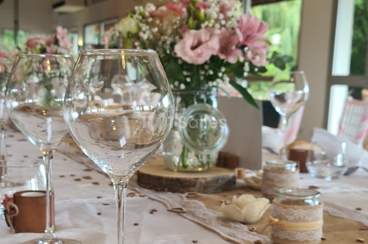 Table décorée avec des verres à pied, des fleurs roses et blanches, et des éléments de décoration en bois et dentelle.