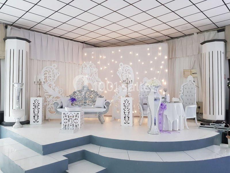 Décor de scène de mariage avec fauteuils ornés, colonnes blanches, guirlandes lumineuses et table dressée.