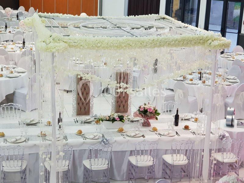 Table de réception élégamment décorée avec des fleurs blanches, des chaises transparentes et une structure ornée de…