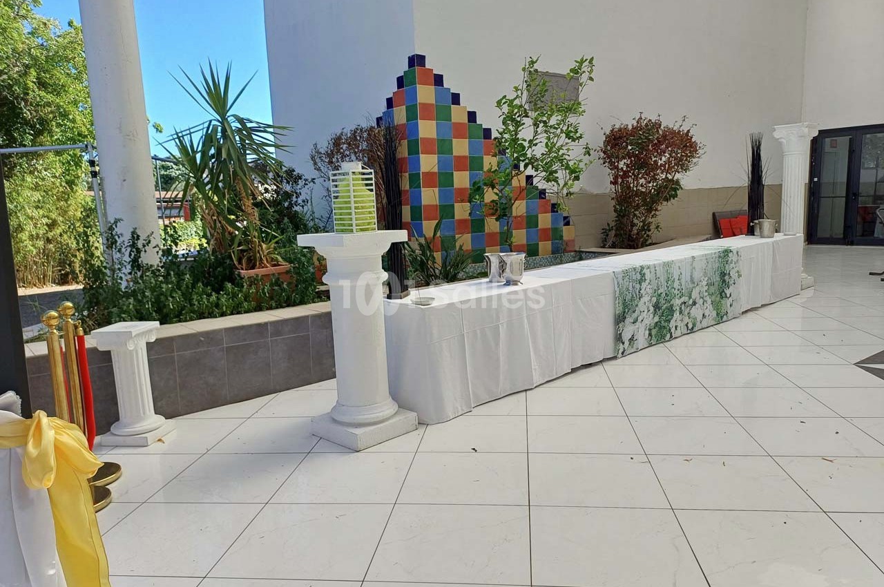 Table drapée de blanc avec colonnes décoratives, située dans un espace extérieur carrelé et entourée de verdure.