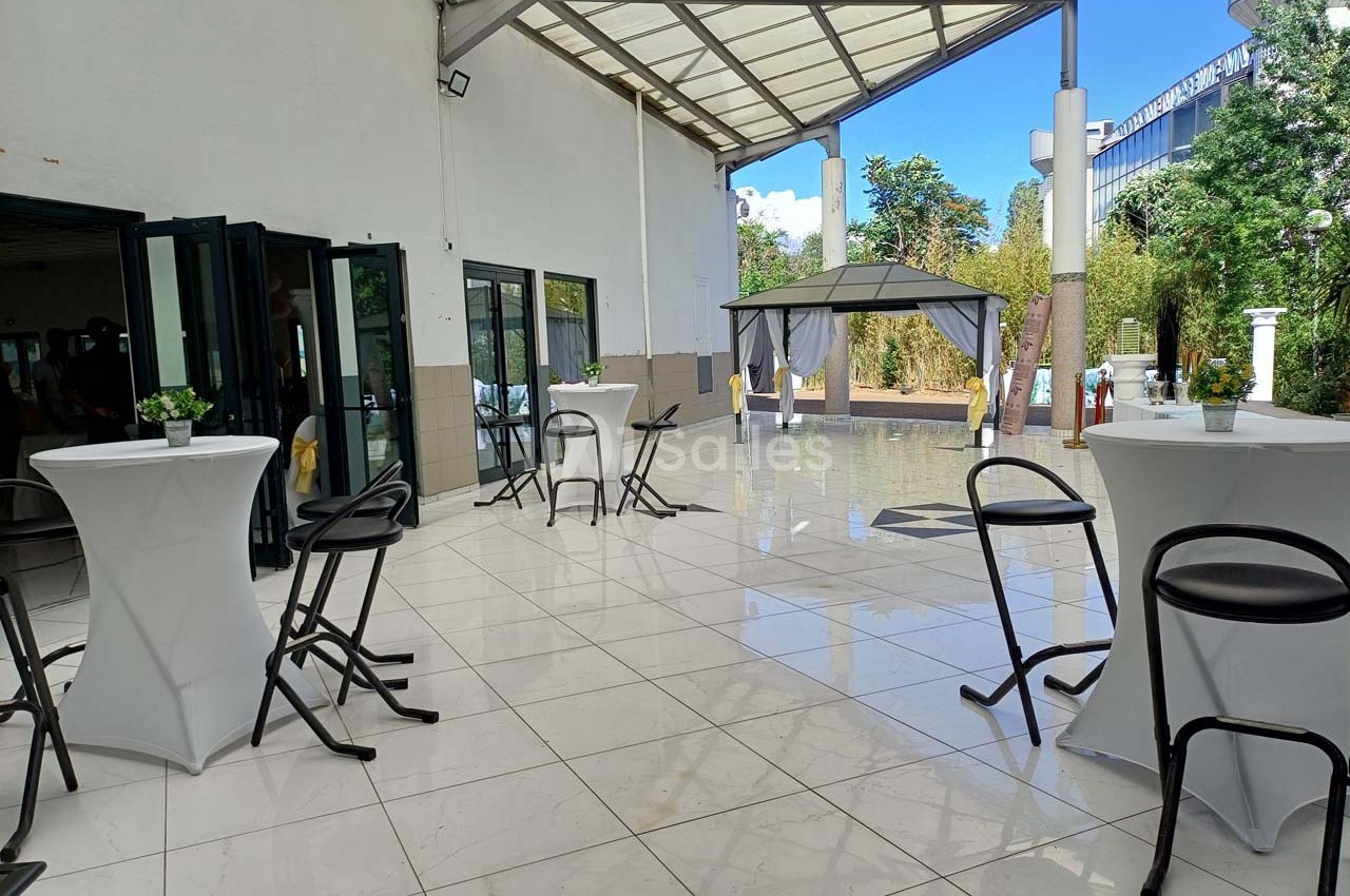 Terrasse couverte avec sol en carrelage blanc, tables hautes, chaises noires et tonnelle dans un espace extérieur arboré.