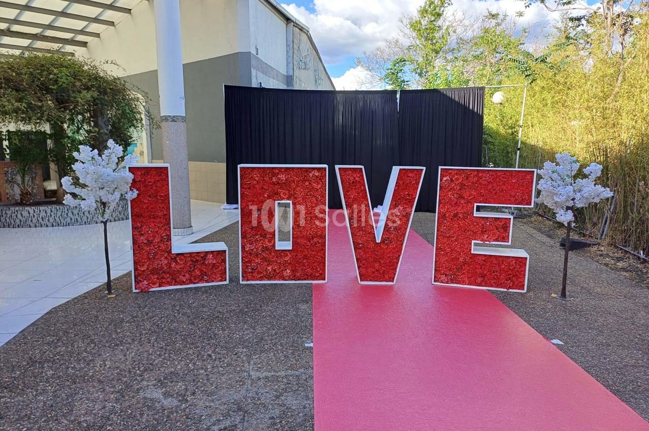 Lettres rouges formant le mot ’LOVE’ sur un tapis rouge, entourées de petites décorations blanches, en extérieur.
