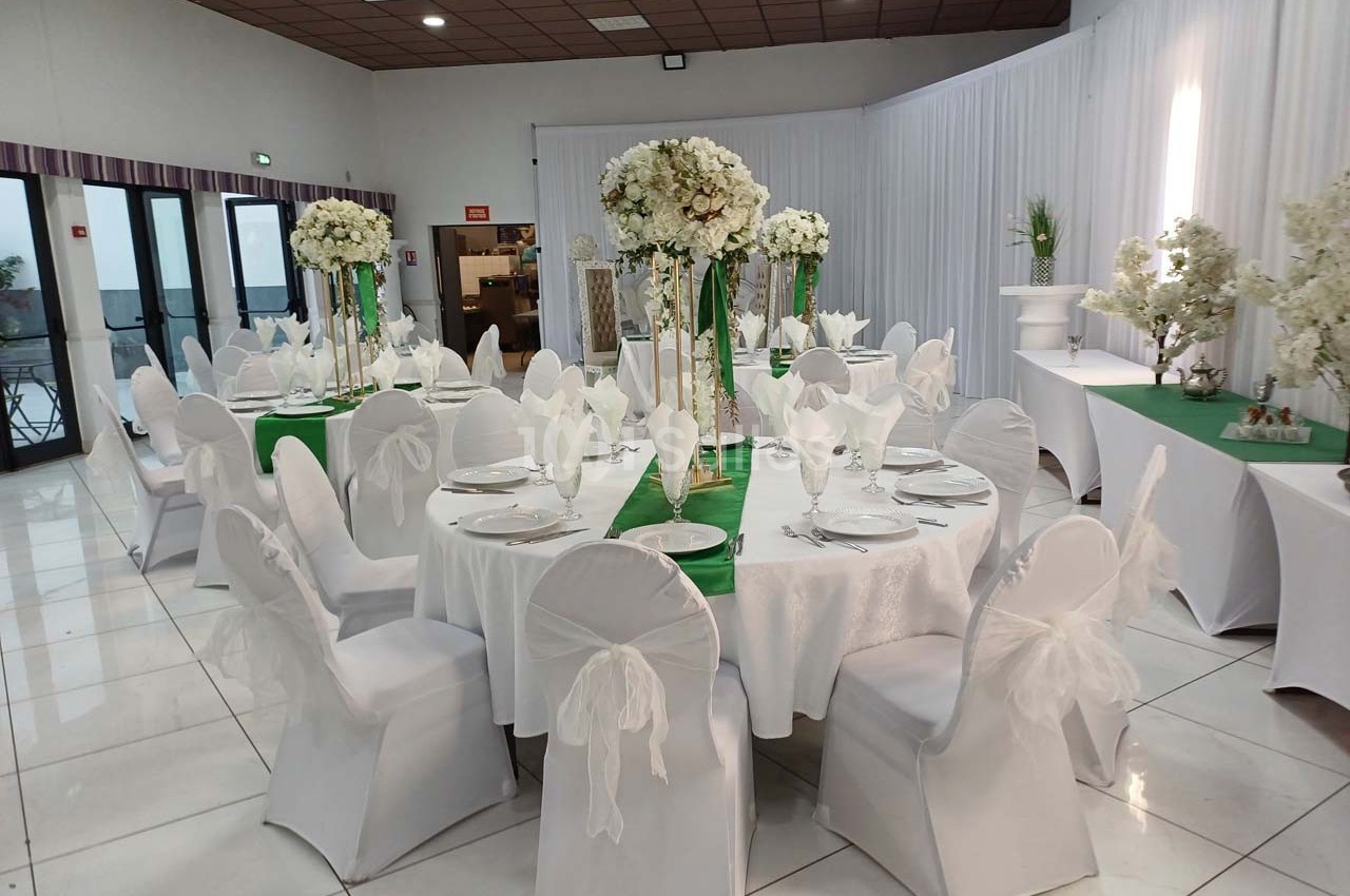 Salle de réception décorée avec des tables rondes, nappes blanches, chaises habillées et centres de table fleuris.